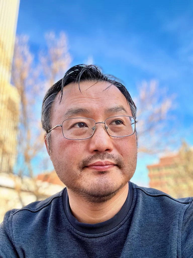 Picture of Wyatt who is Asian American and wearing glasses. Wyatt is shown from the shoulders up wearing a dark gray sweater. Background is a blurry downtown cityscape and sky.